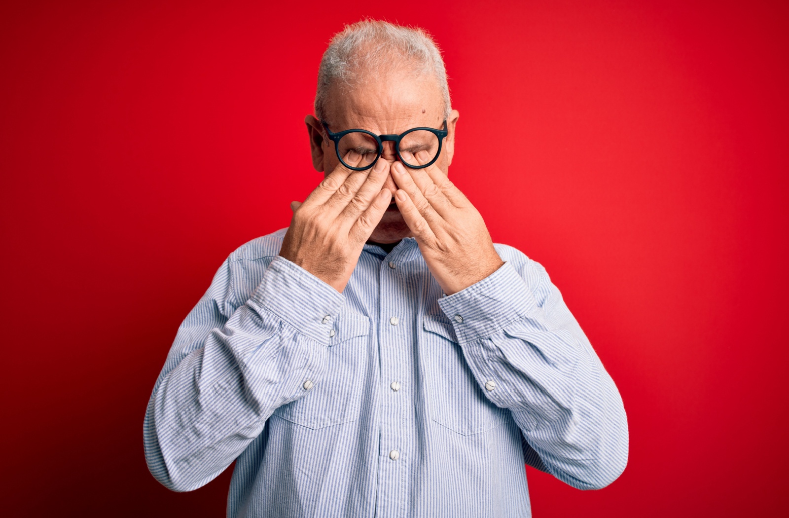 A mature adult rubbing their eyes under their glasses against a bright red background.