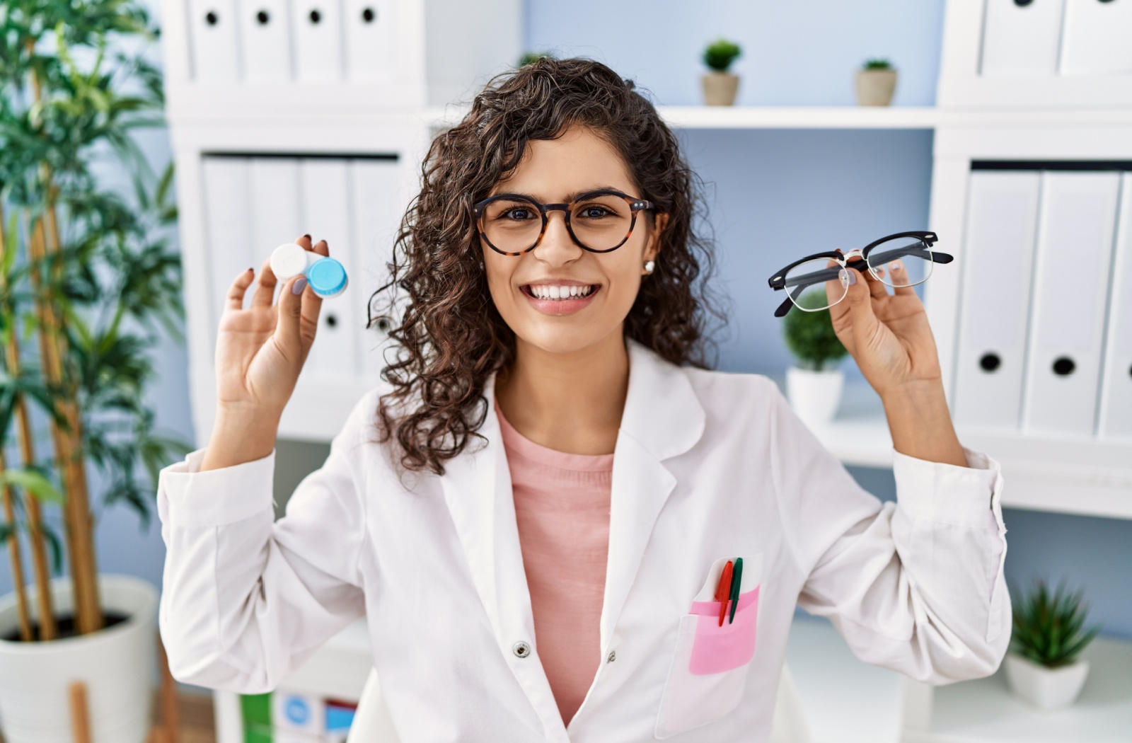 A young optometrist holding a pair of glasses in one hand and a contact lens case in the other.