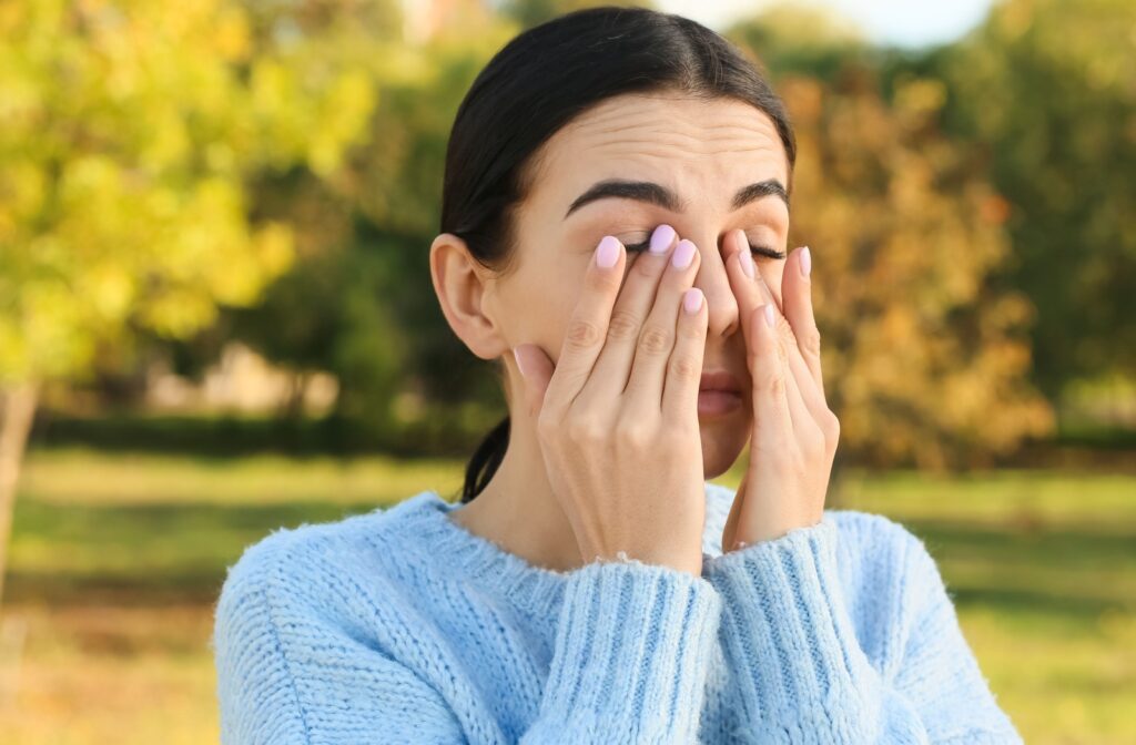 Woman with dry eyes standing outdoors with eyes closed, gently pressing her fingers against both eyelids.