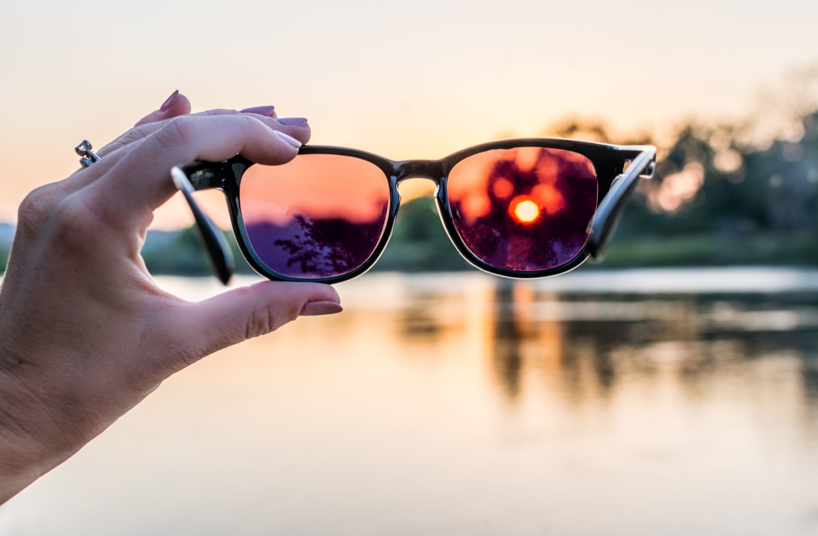 Person holding sunglasses at sunset over a lake.