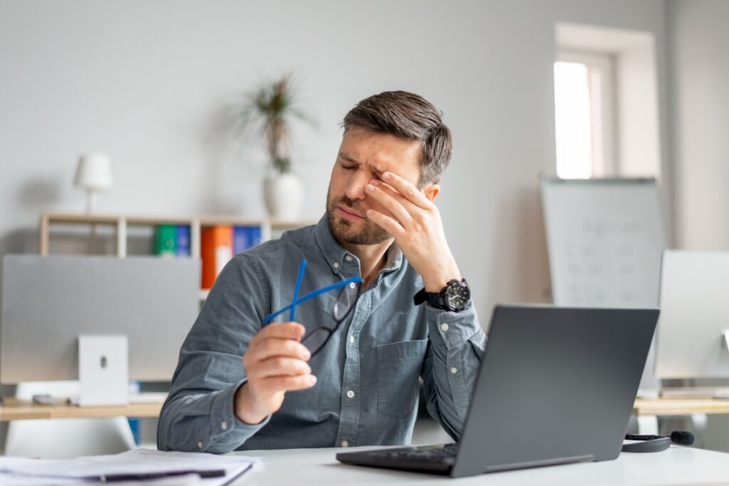 Man at office desk rubbing his eyes and holding eyeglasses while experiencing eye fatigue from prolonged laptop computer use.