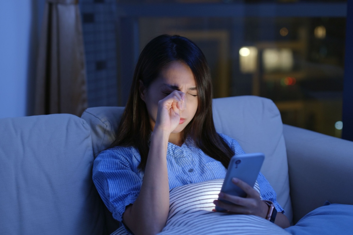 Woman rubbing her eyes while using smartphone on couch at night experiencing digital eye strain and fatigue from blue light exposure.