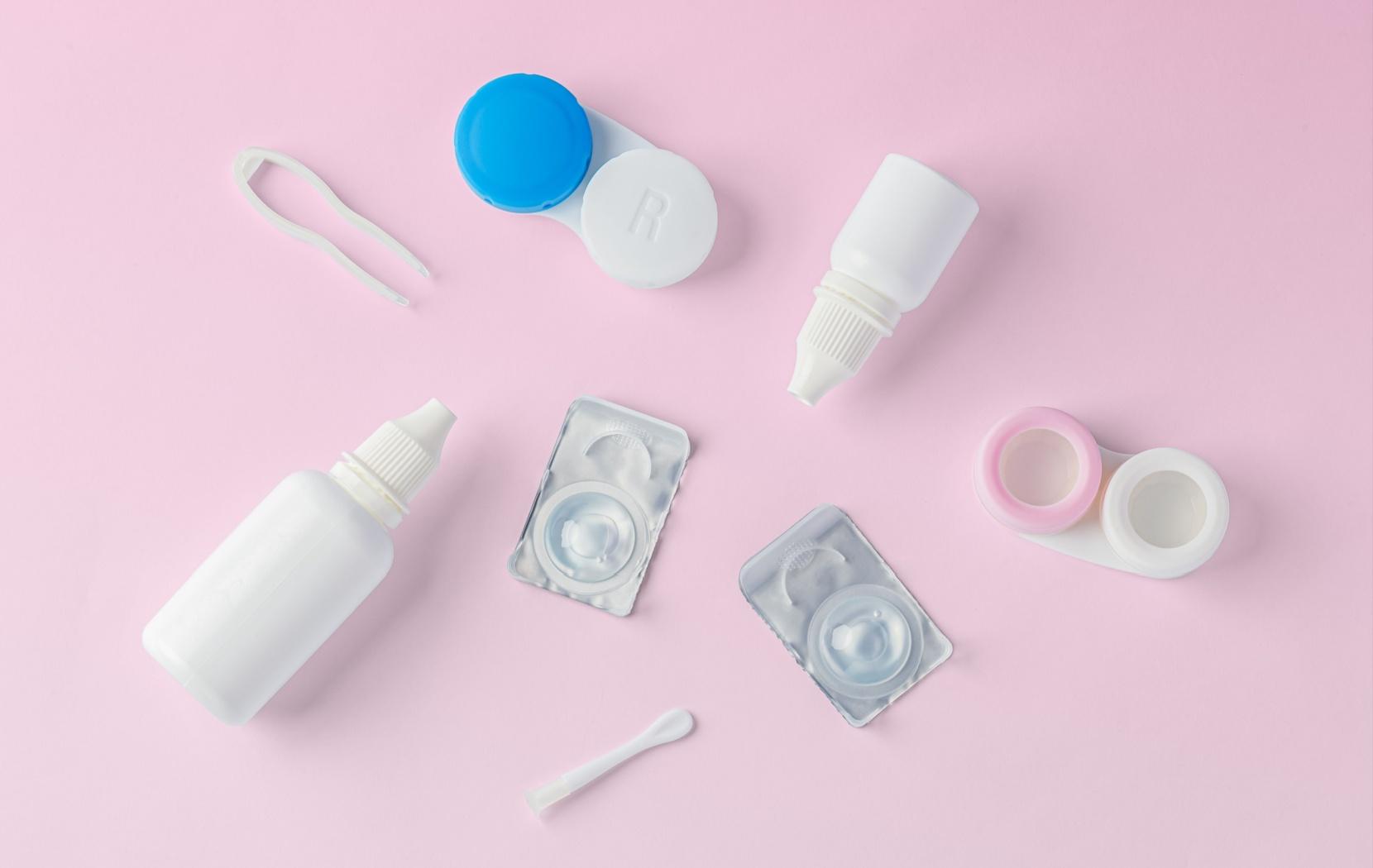 Contact lens supplies including cases, solution bottles, and packaged lenses on a pink background.